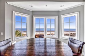 A dining room with a large wooden table and three windows with a beach view. - Oceanfront Estate - Pool, Hot Tub, Pool Table, Bonus Boat- Exclusive Location (Hilton Head Island)