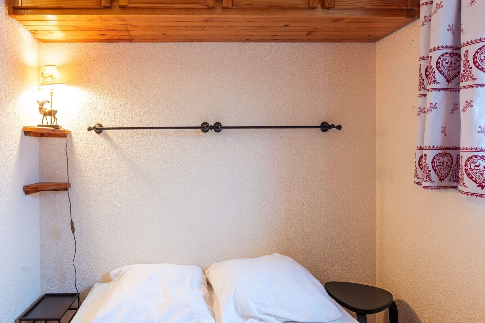 A bedroom with a bed, a bedside table with a lamp, a wall-mounted shelf, and a curtain with a heart pattern.