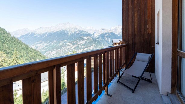 A balcony with a wooden railing, a single lounge chair, and a view of snow-capped mountains.