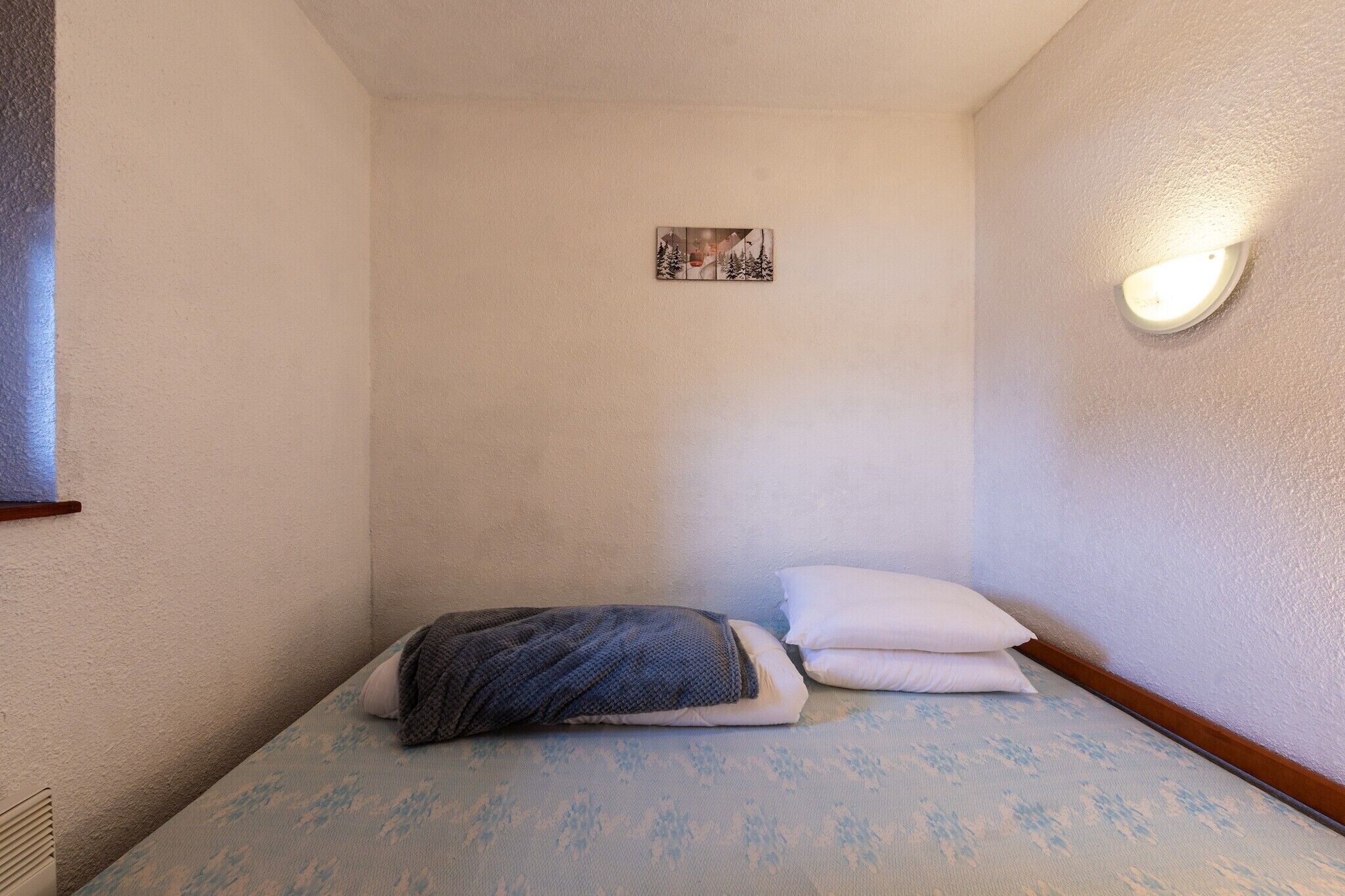 Una habitación pequeña y acogedora con una cama, un almohadón, una lámpara de pared y una fotografía.