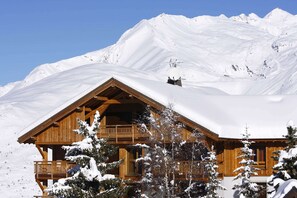 A wooden chalet with a balcony, surrounded by snow-covered trees and mountains.