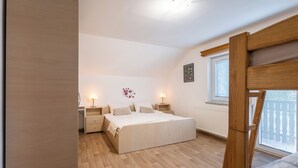A bedroom with a wooden floor, a bed with a white bedspread, two bedside tables with lamps, a window with blinds, and a wooden ladder. - Mountain Nature Apartment with Balcony in Savinja Valley (Mozirje)