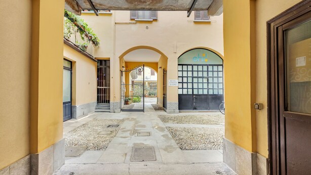 A narrow alleyway with a yellow wall, a metal gate, and a door on the right. - Naviglio 13 - Welcomely (Milan)
