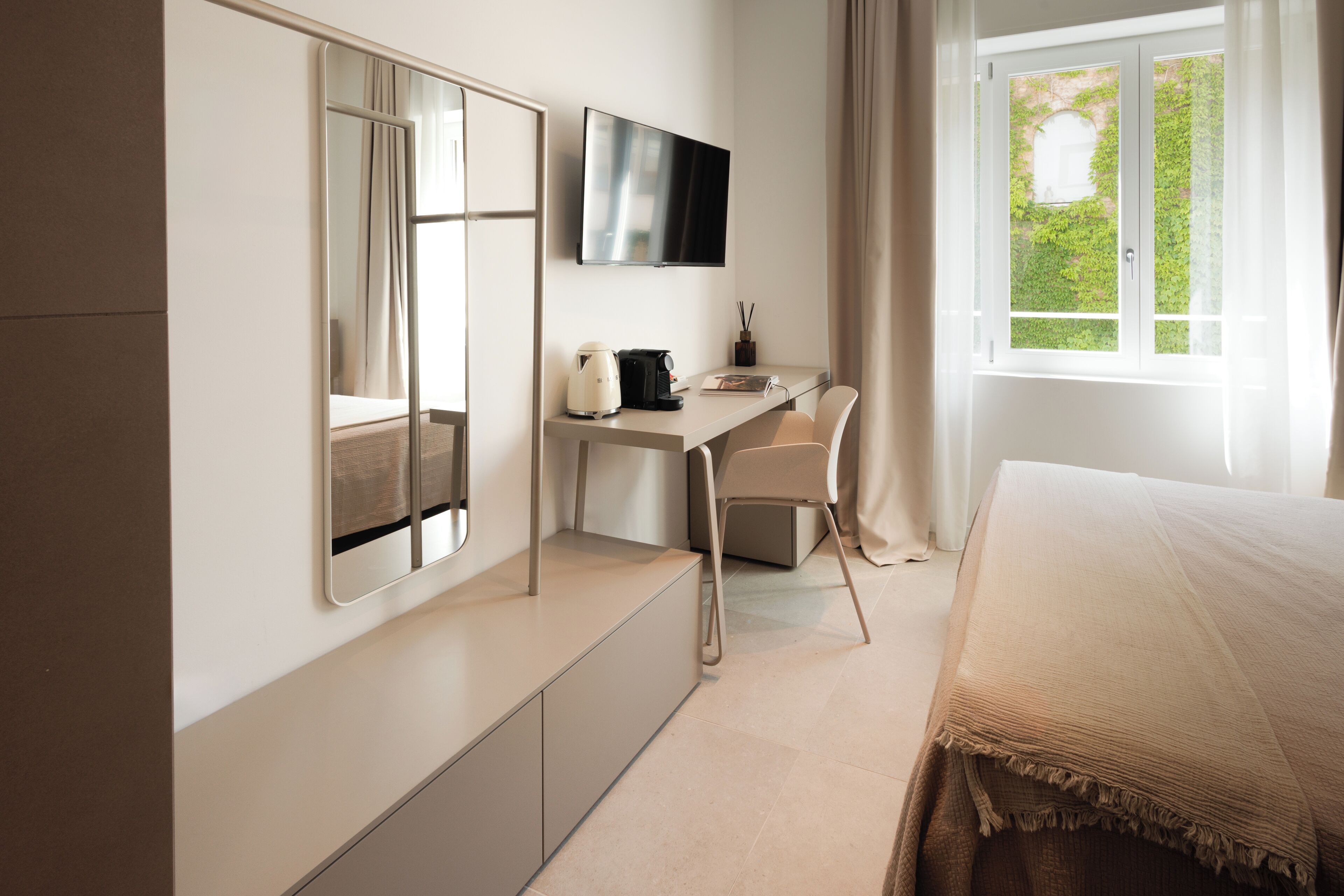 A modern hotel room with a bed, a desk with a chair, a television, and a window with curtains.