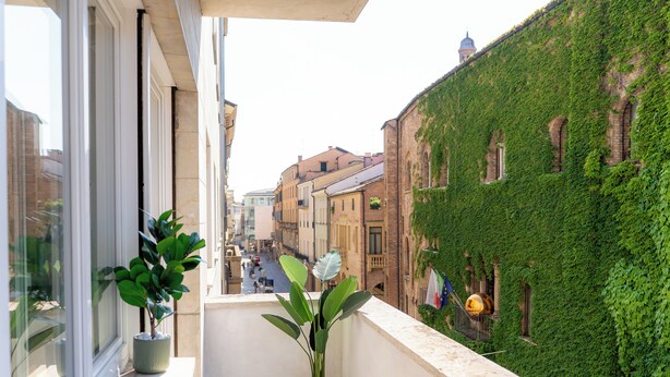 Un balcón con macetas, con vista a una calle con edificios históricos y un edificio cubierto de hiedra.