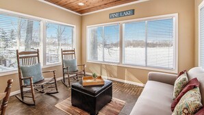 Un salon chaleureux avec un plafond en bois, de grandes fenêtres équipées de stores, un fauteuil à bascule, un canapé et une pancarte sur laquelle on peut lire « Pine Lake ».