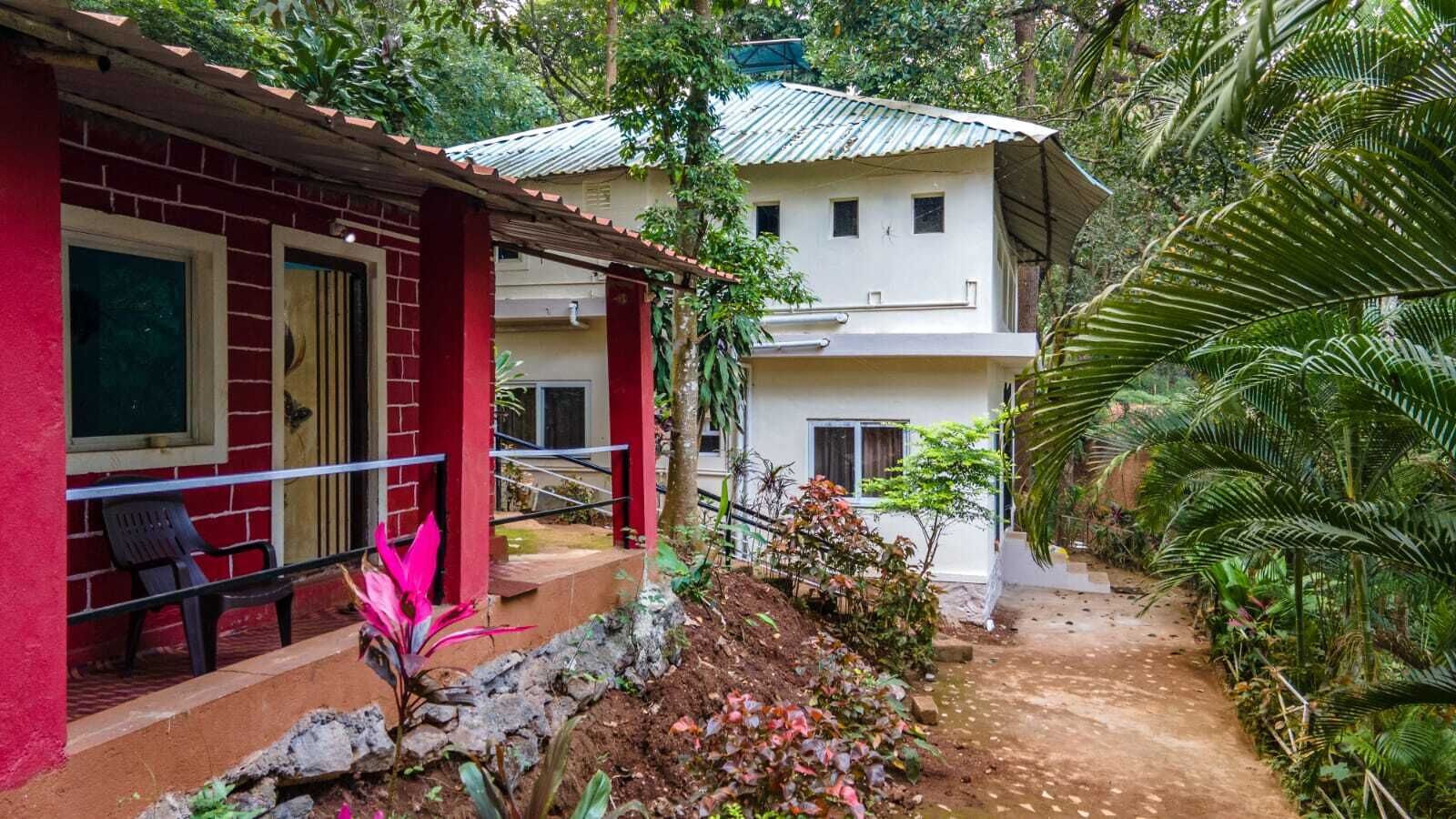 Un bâtiment à deux étages, à la façade rouge et au niveau supérieur blanc, entouré d’une végétation luxuriante et bordé d’un chemin de terre.