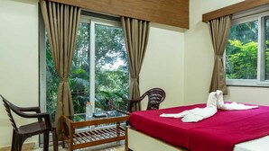 Una habitación de hotel con una cama, una silla de madera, una mesita pequeña y ventanas grandes con cortinas.