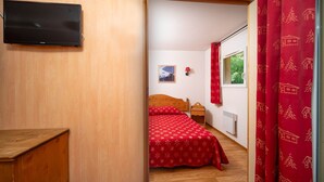 A bedroom with a wooden floor, a bed with a red patterned bedspread, a nightstand, a wall-mounted TV, and a window with red curtains. - Apartment in Alps near Ski Slopes (Allos)