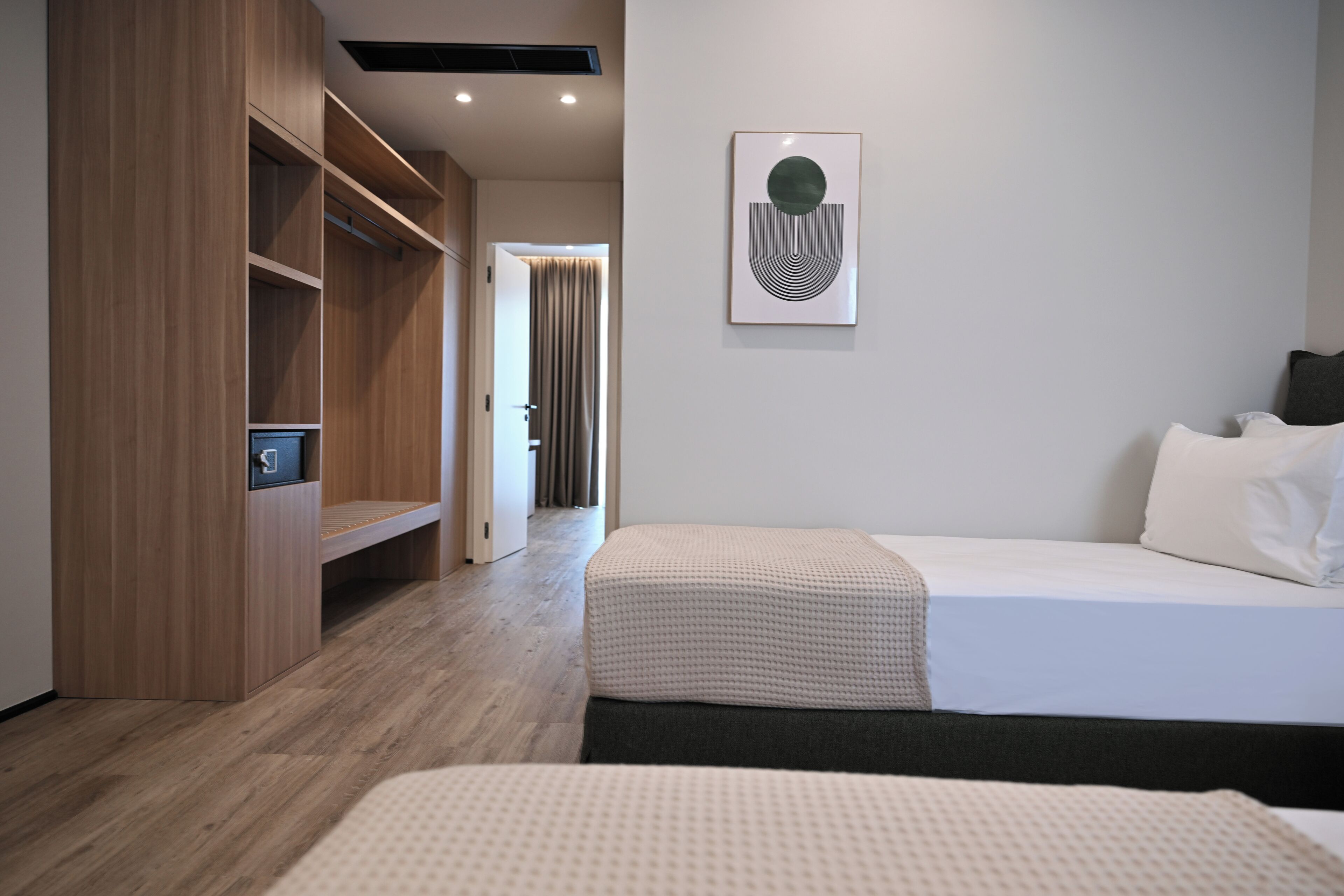 A modern hotel room with a wooden wardrobe, a bed with a white mattress and dark headboard, a framed abstract artwork on the wall, and a wooden floor.
