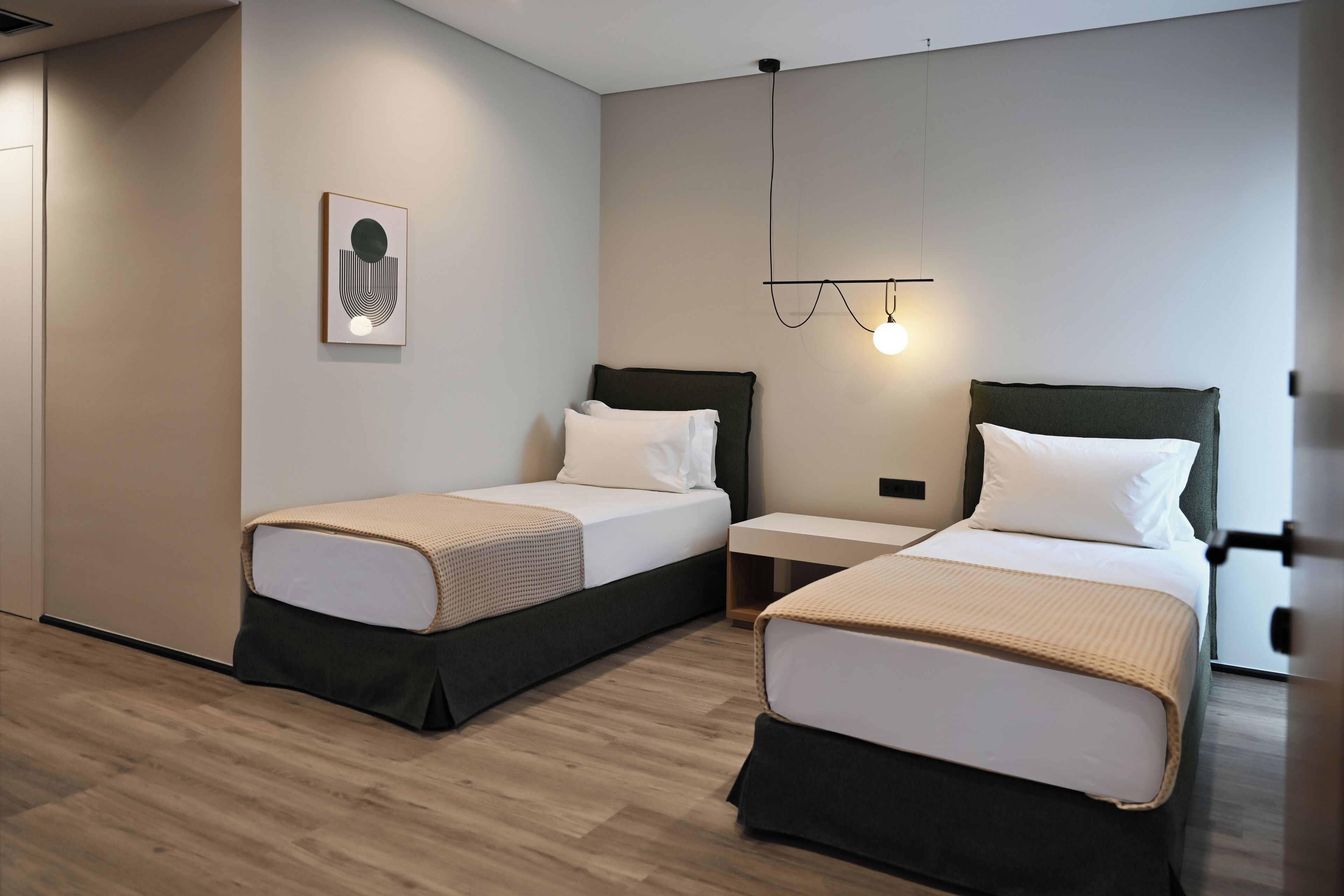 A modern hotel room with two beds, a bedside table, a wall-mounted light, and a framed picture.