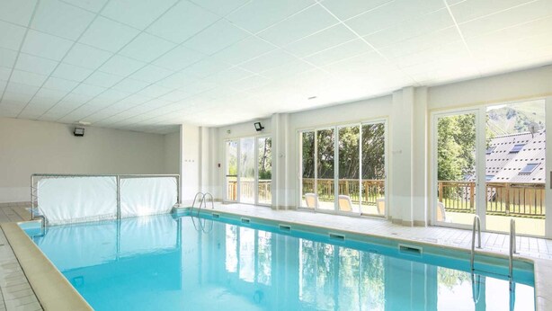 An indoor swimming pool with clear glass walls, a tiled floor, and a view of a residential area with trees and a house. - Apartment in Saint-Sorlin near Ski Resort (Saint-Sorlin-d´Arves)