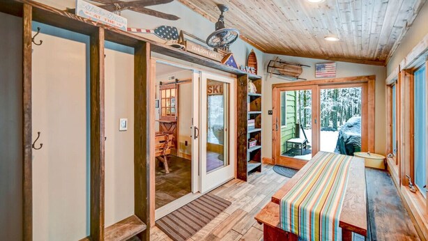 A rustic cabin interior with a wooden ceiling, a dining table with a striped tablecloth, and a view of a snowy landscape through the door.