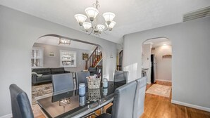 Un coin repas avec une table à plateau en verre, des chaises à revêtement gris, un lustre et une vue sur le salon.