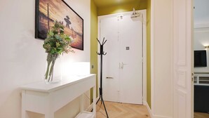 En hall med ett vitt konsolbord, en vas med gröna växter, en inramad tavla, en klädhängare och en stängd dörr.