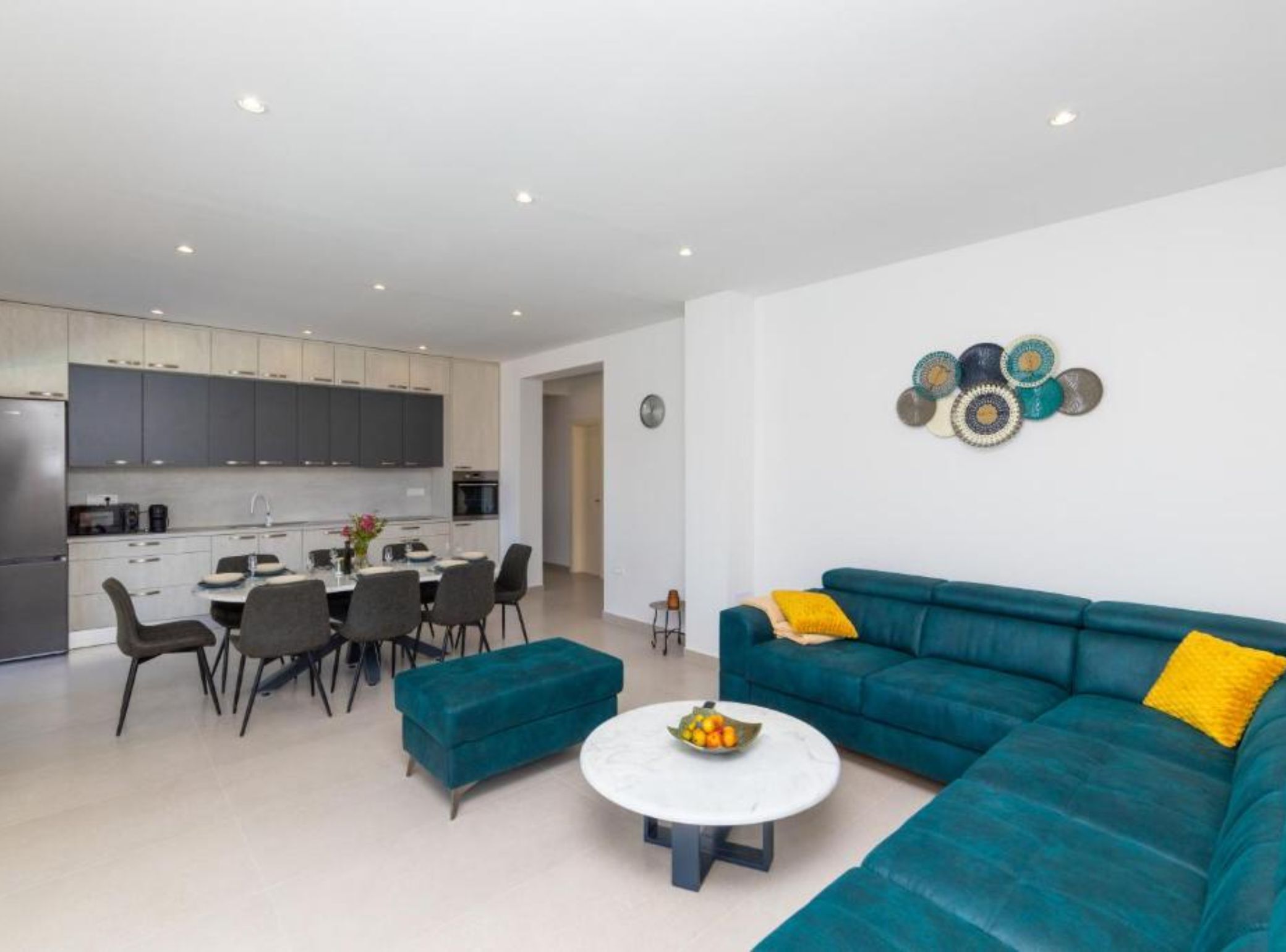 Uma sala de estar moderna com um sofá azul-esverdeado, uma mesa de centro branca e uma área de jantar, com uma cozinha ao fundo.