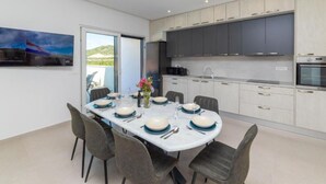 A modern kitchen with grey cabinets, a built-in oven, and a dining area with a round table set for six. - Luxury Villa Navis (Ploce)