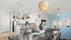 A modern kitchen with white cabinets, a dining area with a dark table, and a living room with a blue wall and a ceiling fan.