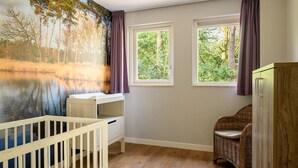 Una habitación con una cuna, un escritorio y una silla de mimbre. Hay dos ventanas con cortinas, y un mural con una escena de bosque en la pared.