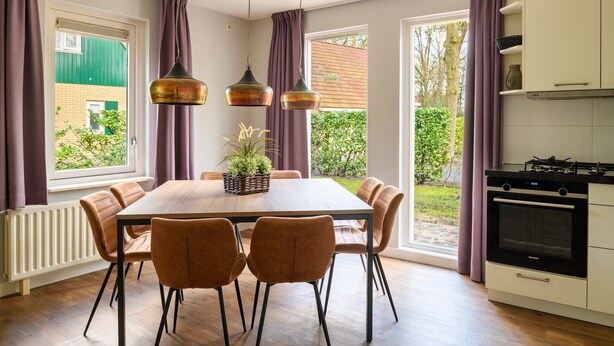 Un comedor con mesa de madera, seis sillas y lámparas colgantes. Se ve una cocina con estufa y horno a la derecha.
