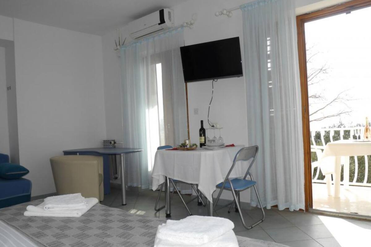 Habitación de hotel con cama, una pequeña mesa con una botella y vasos, un televisor montado en la pared y vistas al exterior por una puerta de vidrio.