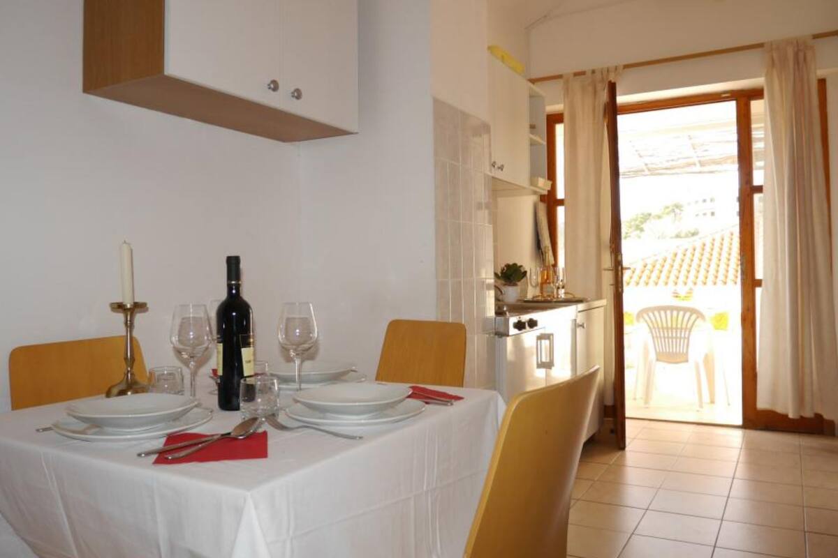 Un comedor con una mesa dispuesta para cuatro, una cocina visible a través de una puerta abierta y vistas al exterior.