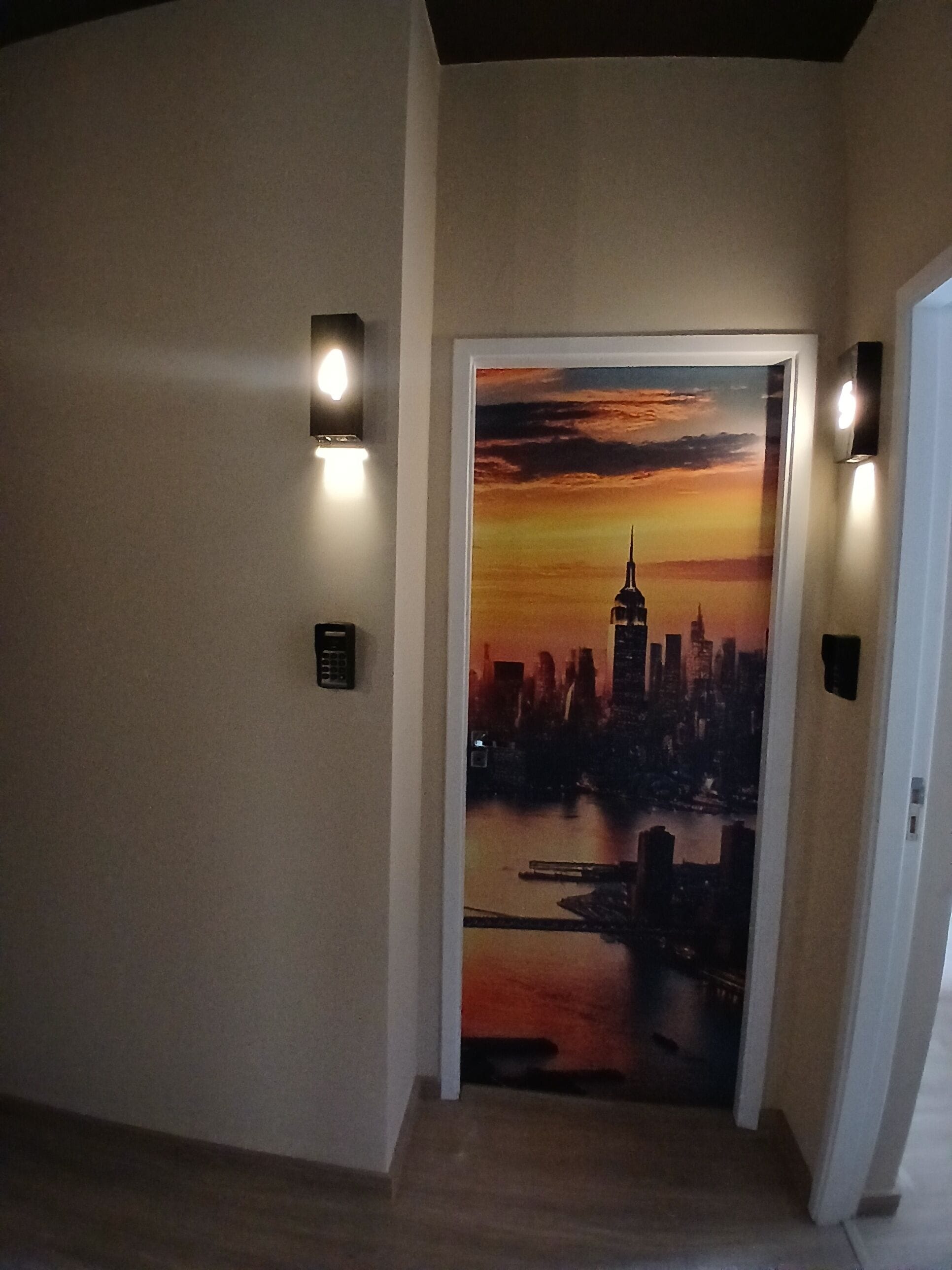 Un couloir avec une porte ornée d’une fresque représentant un paysage urbain au coucher du soleil.