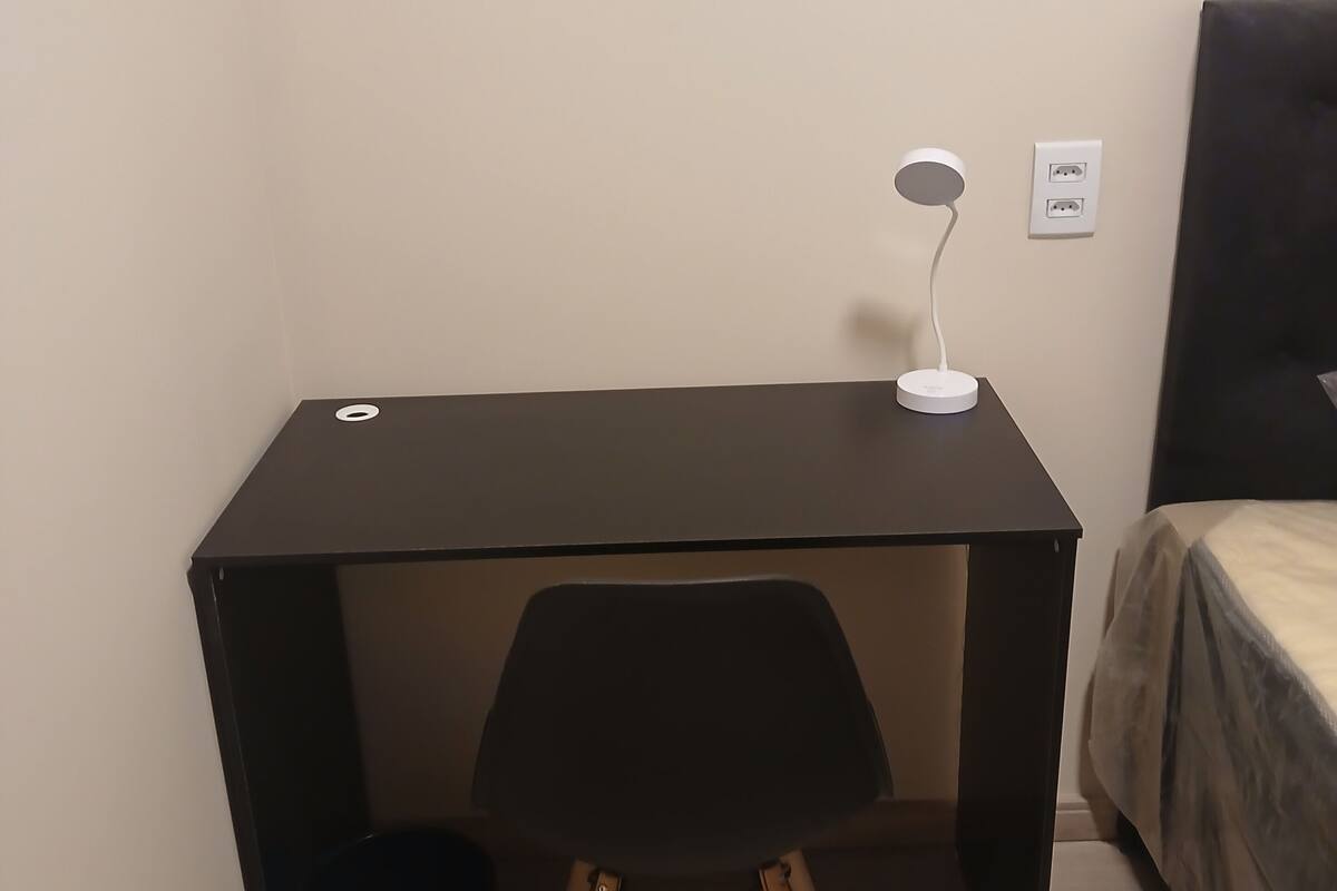 Un petit bureau de couleur sombre, avec une chaise, une lampe de bureau blanche et une poubelle.