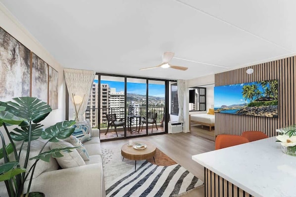 A modern living room with a sofa, coffee table, and a dining area with a white table. There is a balcony with a view of buildings and a ceiling fan.