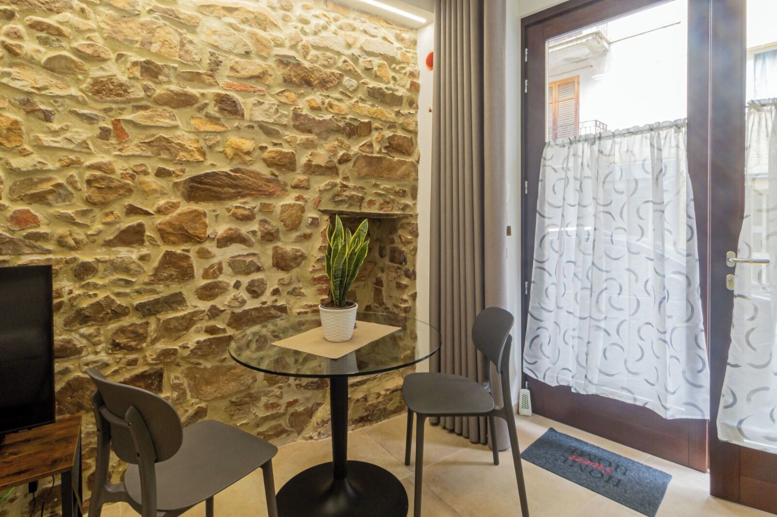 A stone wall, a round glass table with a potted plant, two chairs, a television, and a door with curtains.