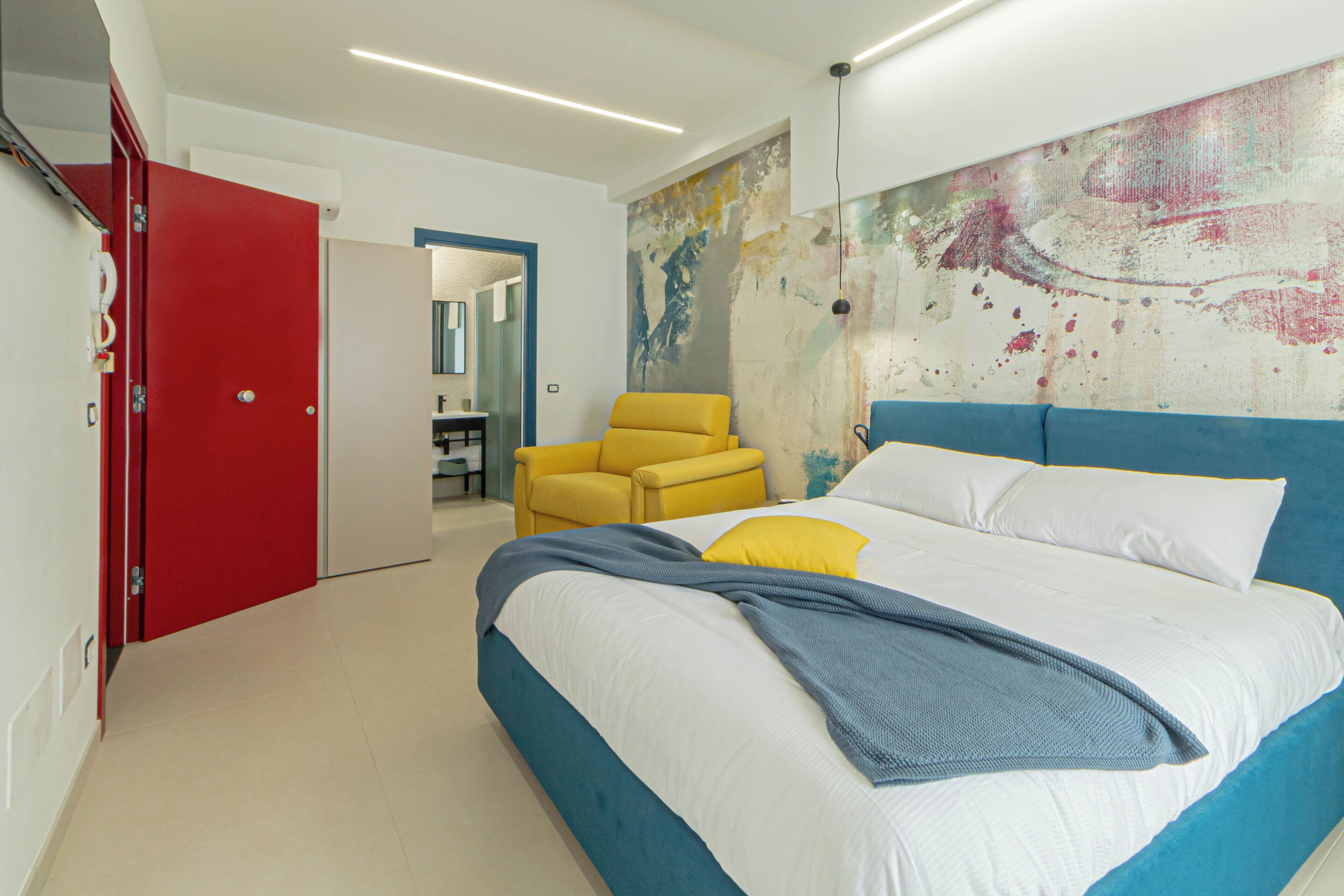 Ein Schlafzimmer mit einem Bett, einem gelben Sessel, einer roten Tür und einer Wand mit einem strukturierten Wandgemälde.