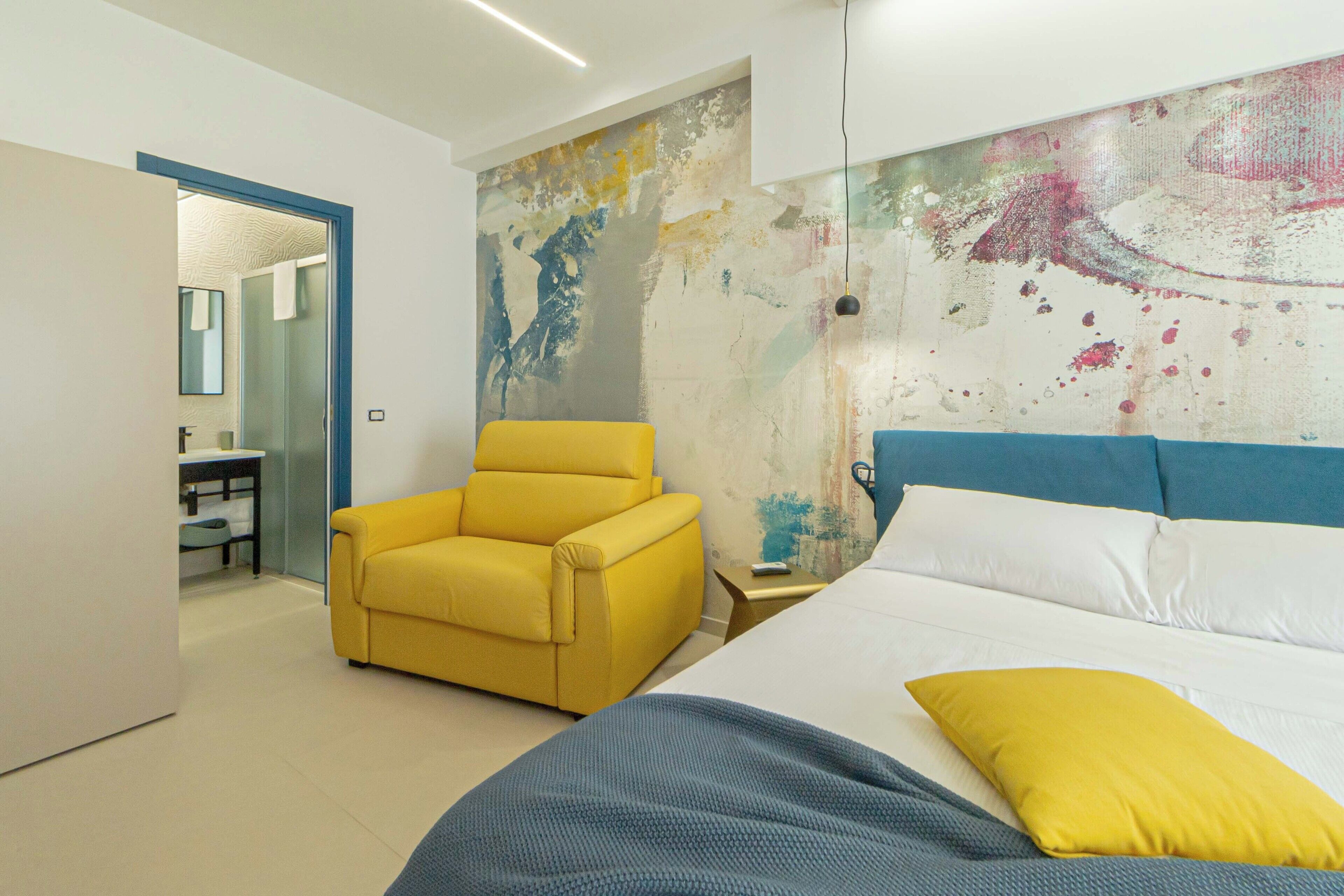 Un dormitorio moderno con cama, un sillón amarillo, un cuadro abstracto grande en la pared y un baño visible a través de una puerta abierta.
