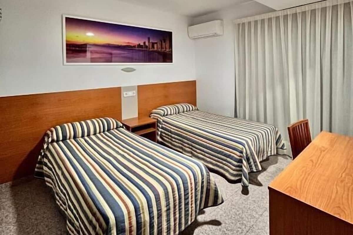 A hotel room with two beds, a wooden desk, a painting of a sunset, an air conditioning unit, and a ceiling light.