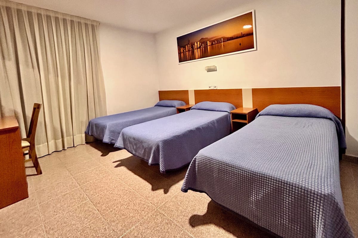 A hotel room with three single beds, a wooden headboard, a desk, and a large window with curtains.