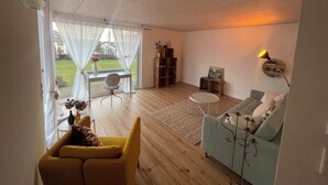 Un salón amplio con suelo de madera, un sillón amarillo, un sofá azul claro, una mesita redonda y una estantería.