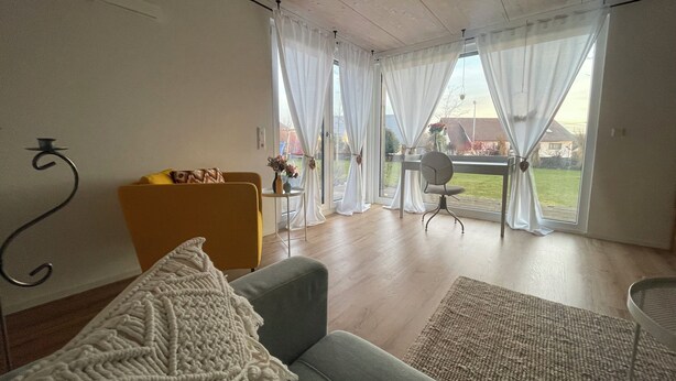 Una sala de estar con un sofá, una mesita con un jarrón, y una silla. Hay ventanas amplias con cortinas, y vistas al exterior.