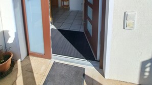 Una entrada con puerta de madera, un pequeño estante de madera con un florero y una planta en maceta afuera.