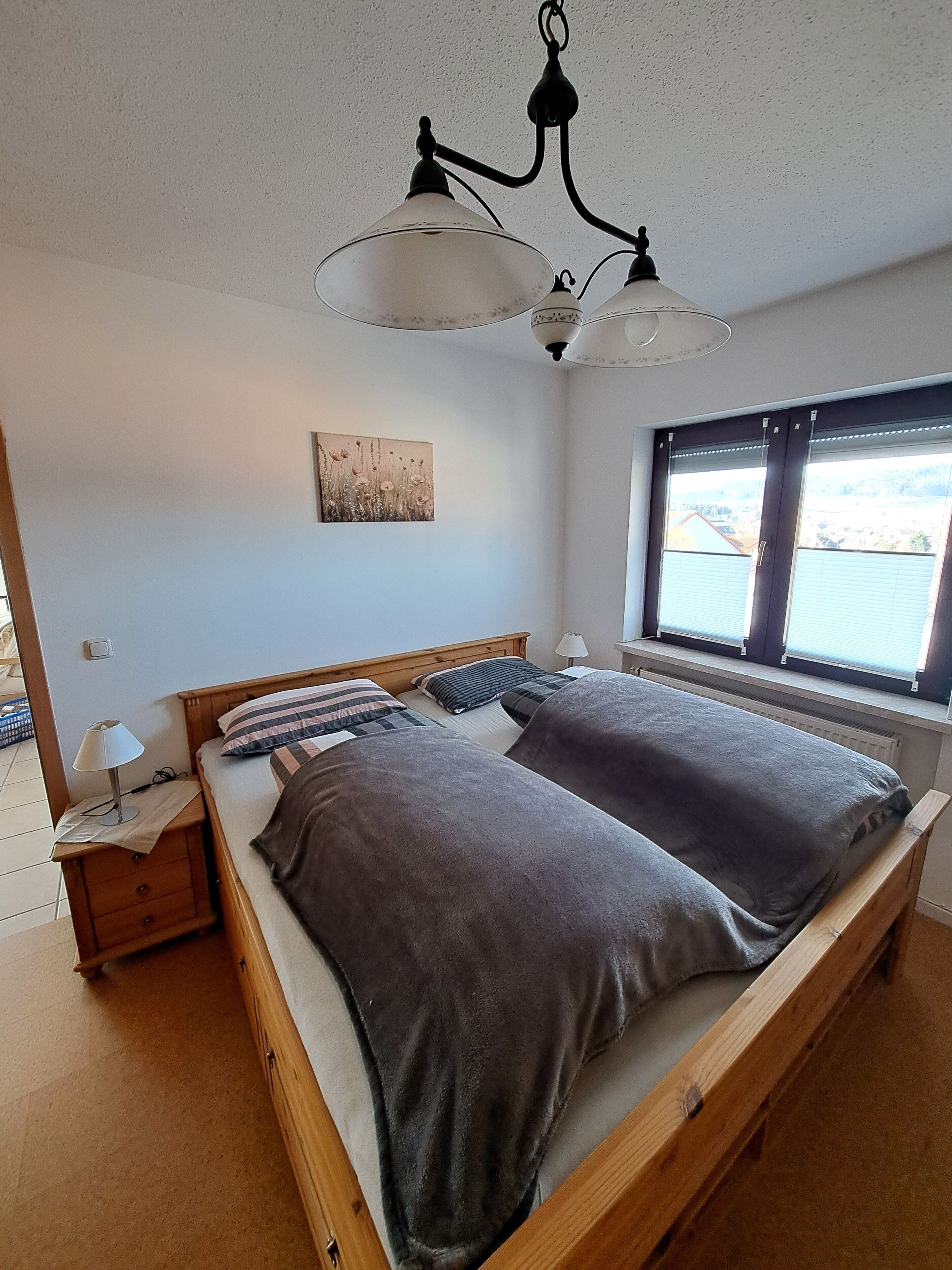 Un dormitorio con cama de madera, una mesita de noche, una lámpara, un aplique en el techo, una ventana con persianas y una obra de arte colgada en la pared.