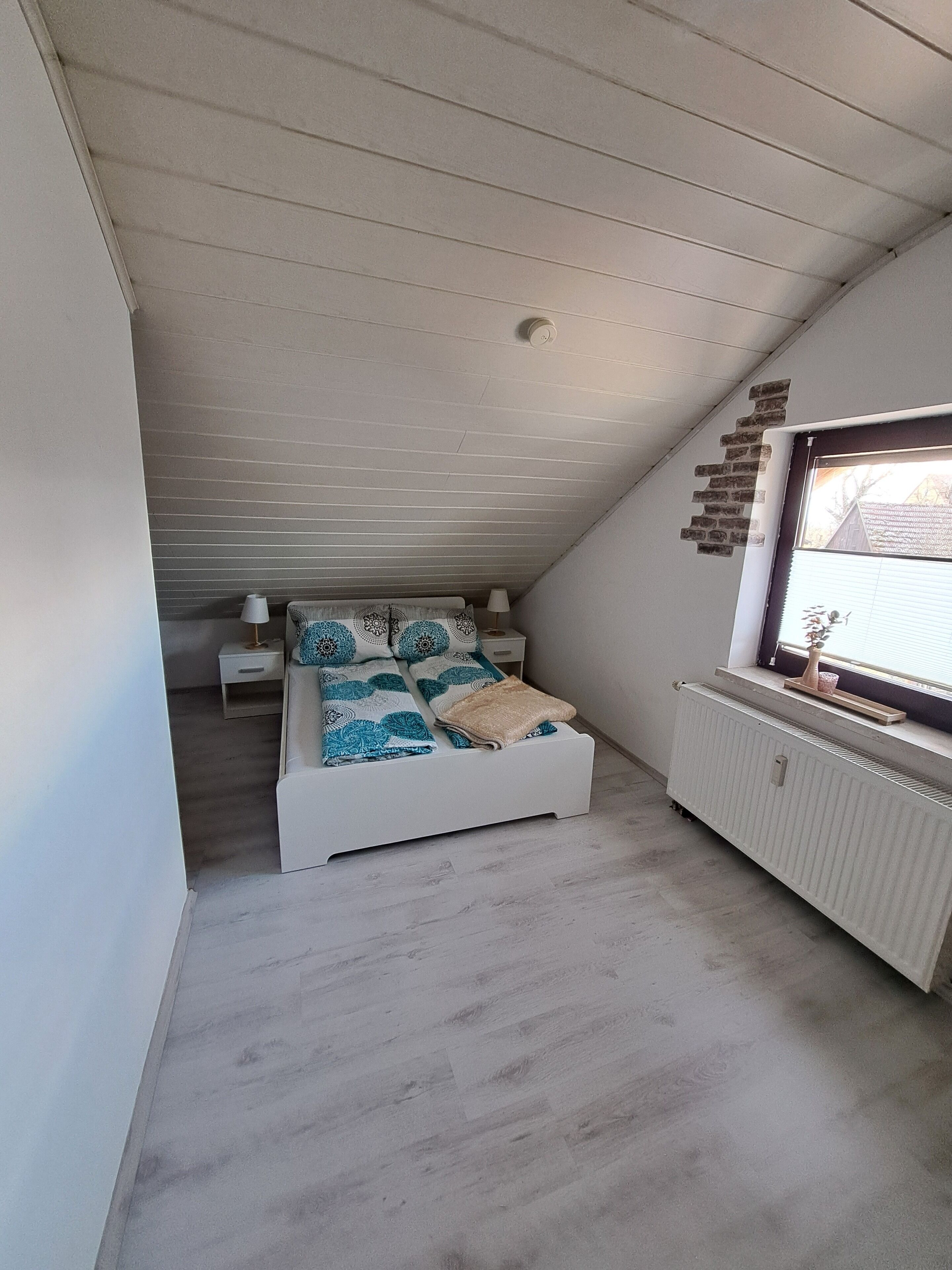 Une chambre avec un plafond en pente, un lit avec une literie à motifs, une table de chevet, une lampe, une fenêtre donnant sur un paysage agréable et un radiateur.