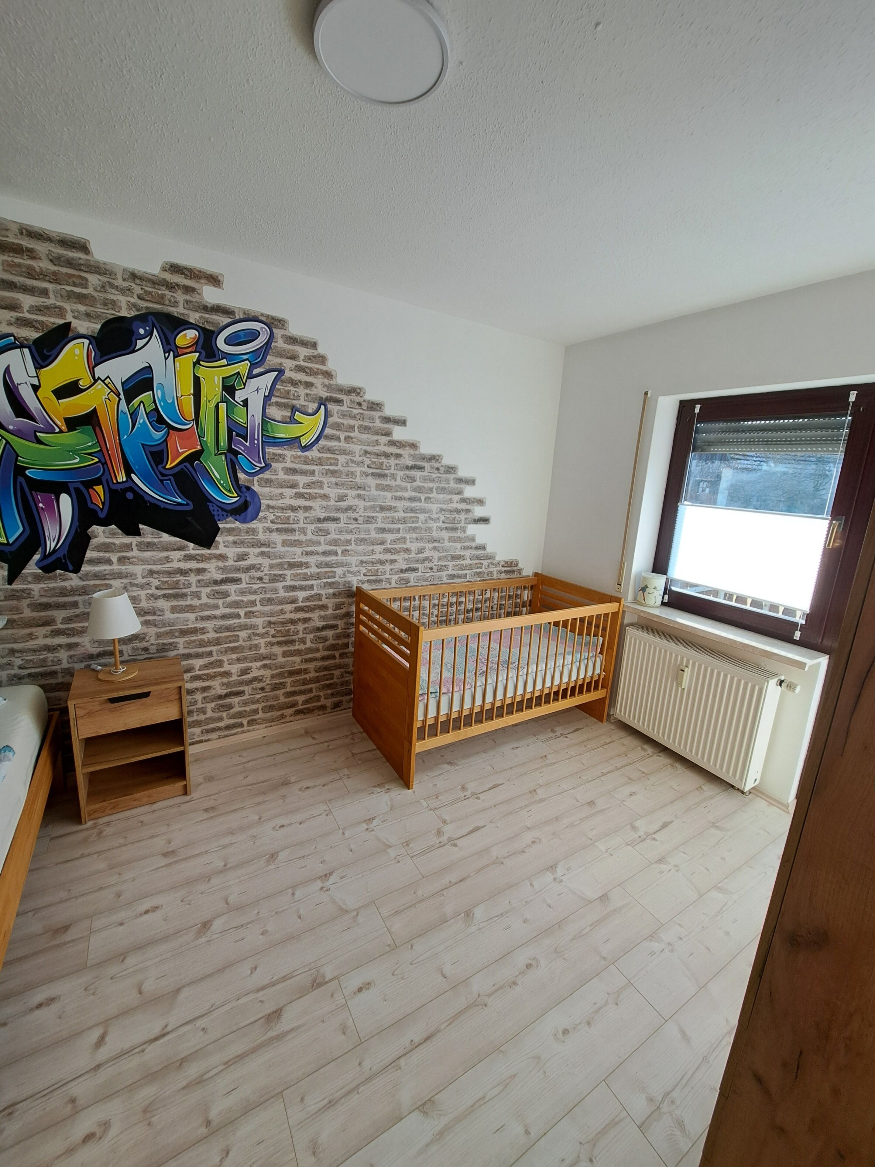 A room with a brick wall, a crib, a wooden nightstand, a radiator, and a window.