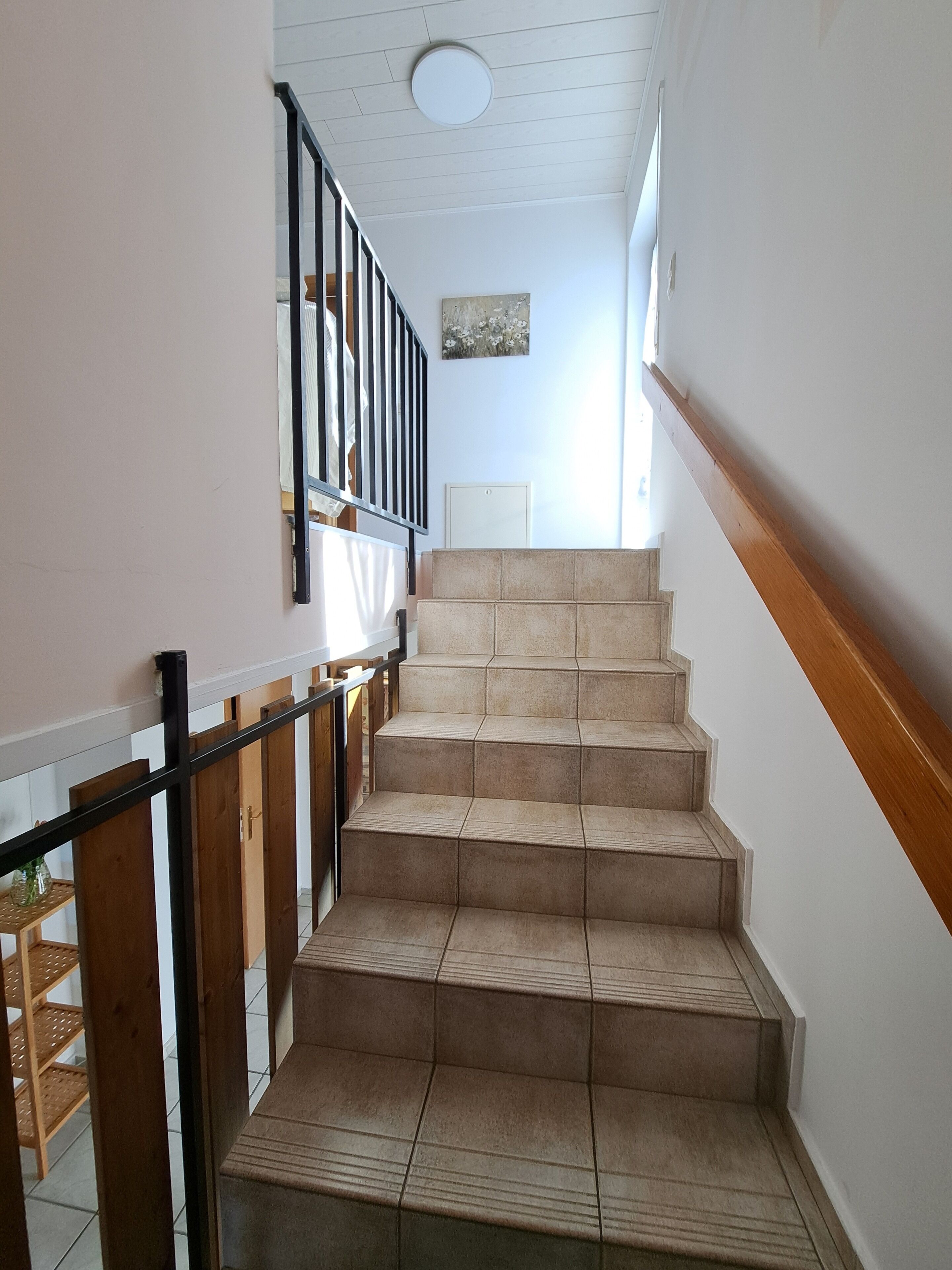 Una escalera con peldaños color beige y una barandilla de madera, que conduce a una plataforma con techo blanco y una lámpara.