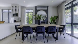 A modern dining area with a large table, six chairs, a central plant, a round mirror, and a built-in air conditioning unit. - Newly Renovated 3 bed Apartment With Splashpool - 2283 (Cape Town)