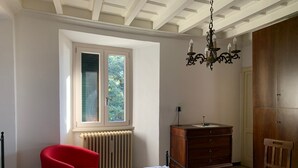 A bedroom with a bed, a red chair, a wooden dresser, a radiator, and a window with a view of trees. - Riflessi Sul Lago (Gravedona)