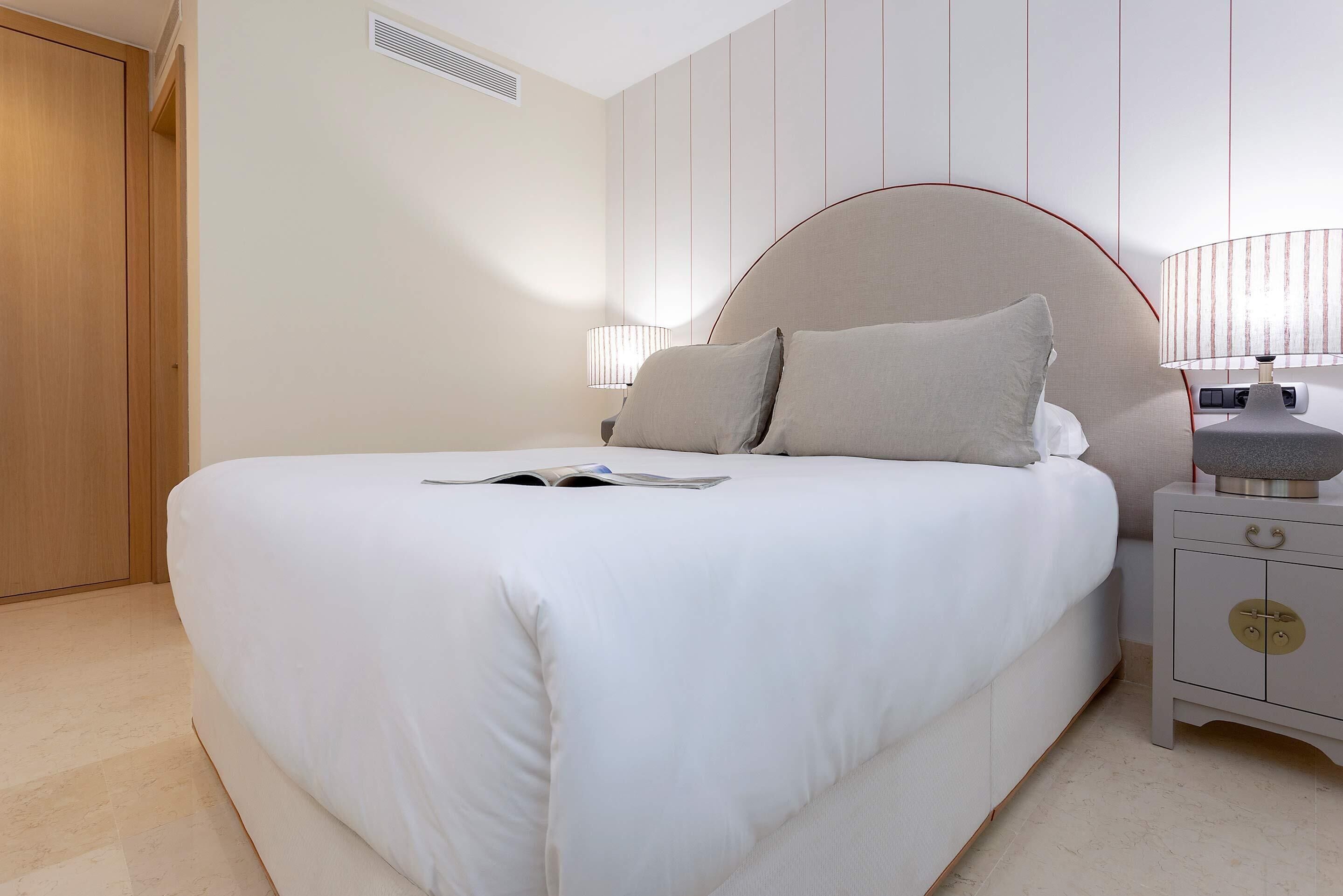 A neatly made bed with a white comforter and pillows, a bedside table with a lamp, and a wooden wardrobe in a hotel room.