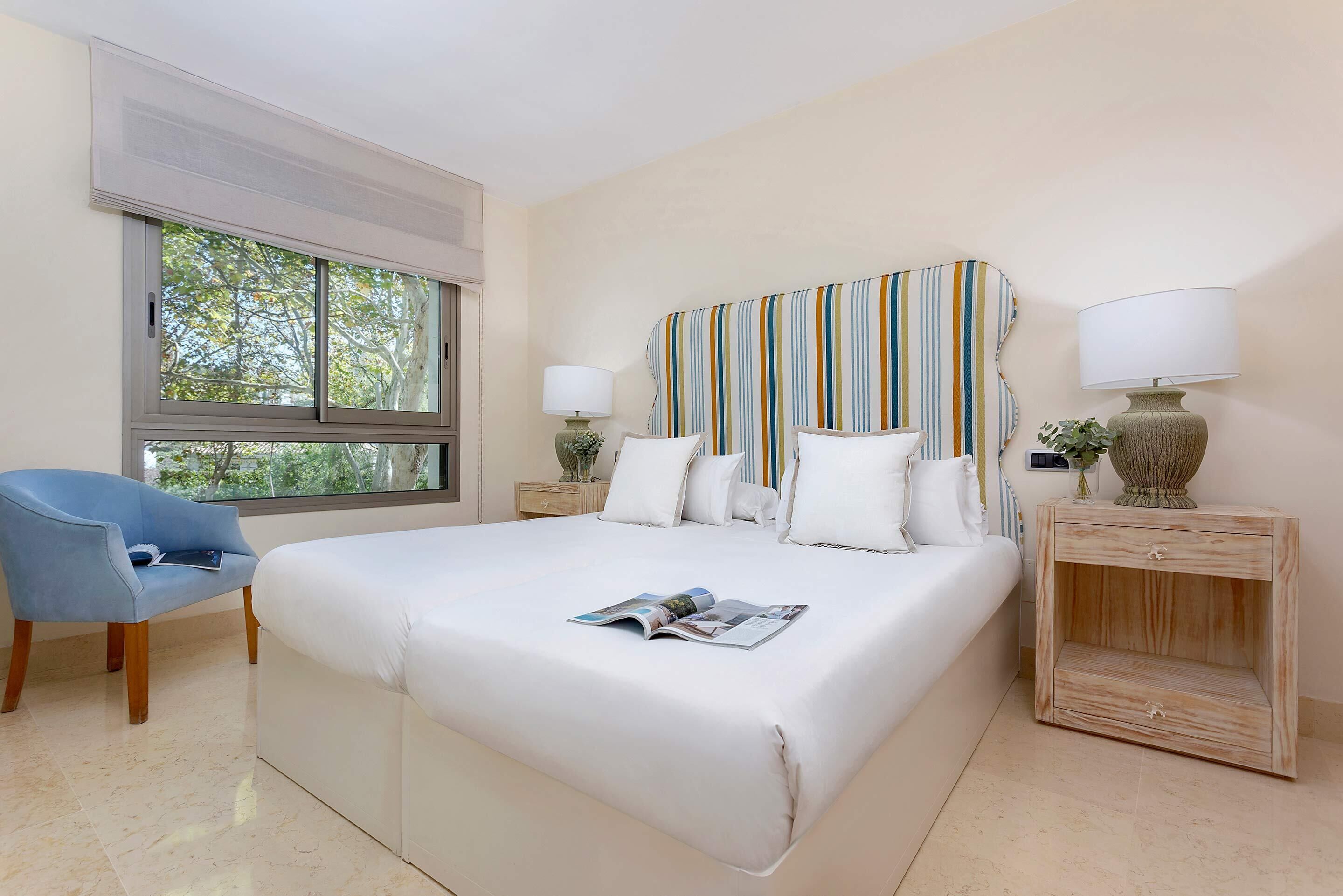A neatly arranged bedroom with a large bed, a blue armchair, a bedside table with a lamp, and a window with a view of trees.