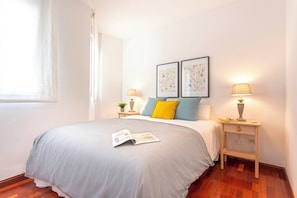 A neatly made bed with a grey bedspread, two pillows, and a magazine on it. There are two bedside tables with lamps and framed pictures on the wall above.