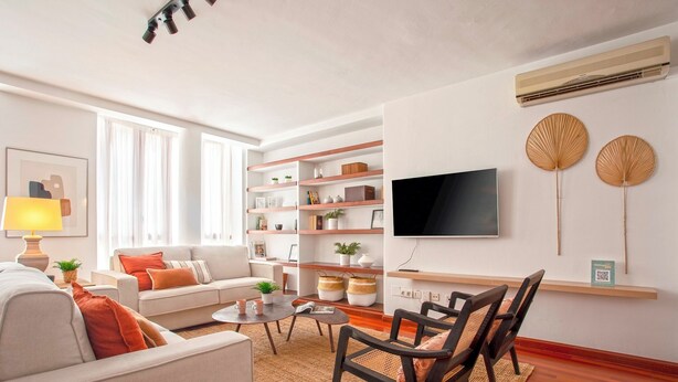 Un salon moderne comprenant un canapé, une table basse et des fauteuils, un téléviseur, des étagères ornées d’objets décoratifs et une grande fenêtre.