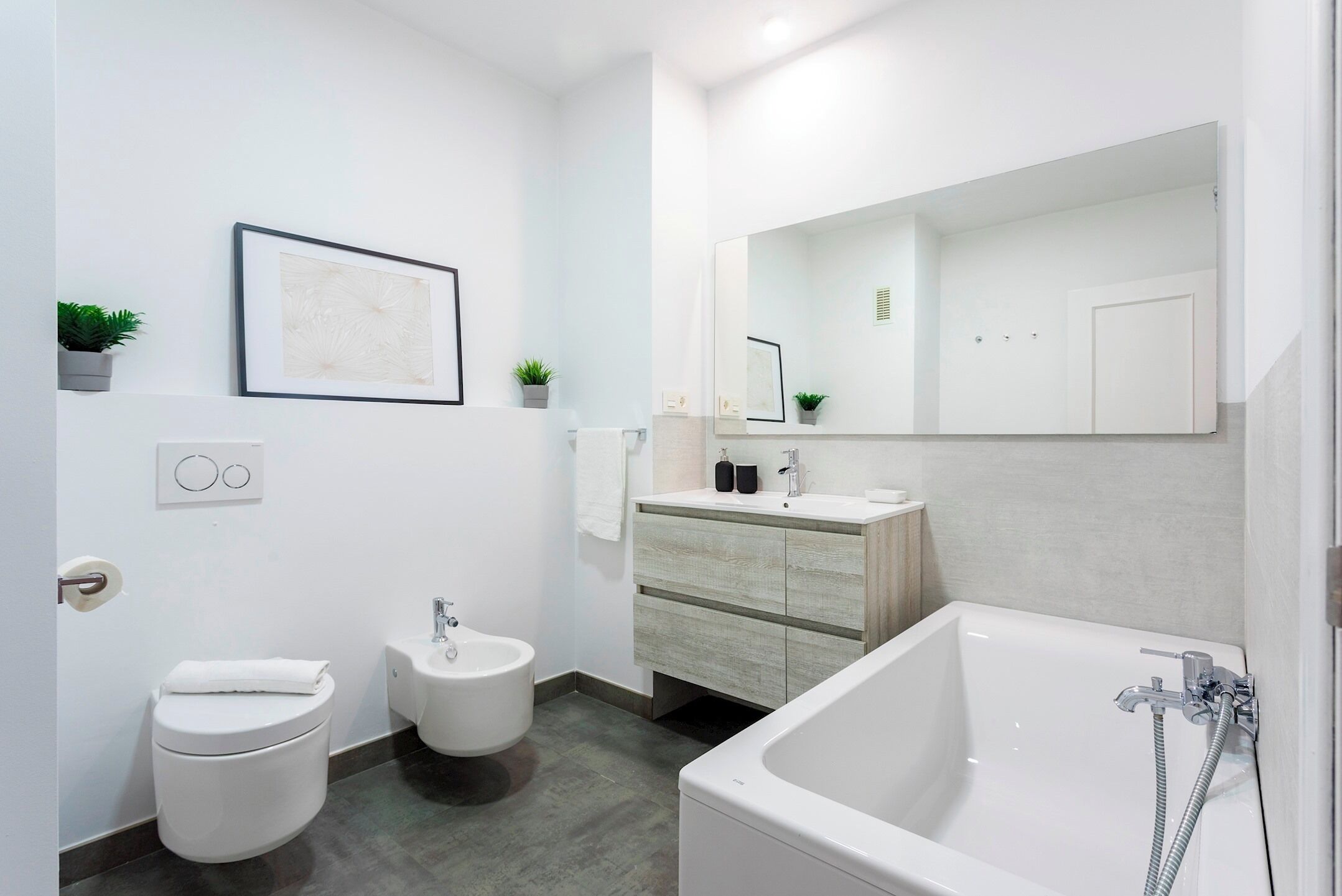 Ein modernes Badezimmer mit Toilette, Bidet, Waschbecken und Badewanne. An den Wänden befinden sich gerahmte Bilder und kleine Topfpflanzen.