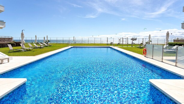 A rectangular swimming pool with clear blue water, surrounded by white tiled edges and a grassy area with lounge chairs and umbrellas. - Genteel Home Guadalmansa Beach (Estepona)