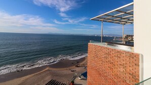 Blick auf einen Strand von einem Balkon mit Glastrennwand, einer Ziegelwand und einem klaren, blauen Himmel.